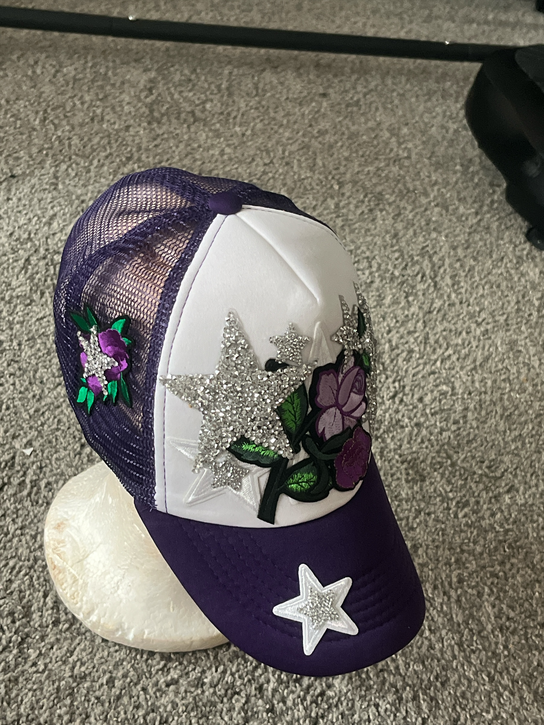Peacefulcornerapparel purple and white trucker hat with embroidered floral motifs and rhinestone stars. Mesh back for breathability.